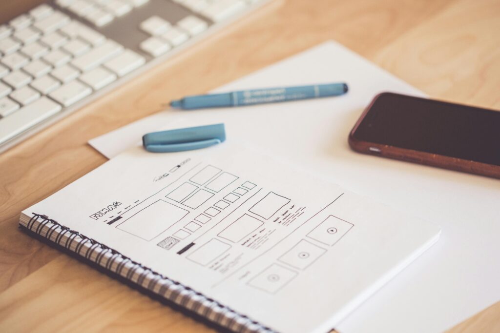 UX/UI Design service A neat workspace featuring a sketchpad with wireframes, a smartphone, and a keyboard on a wooden desk.